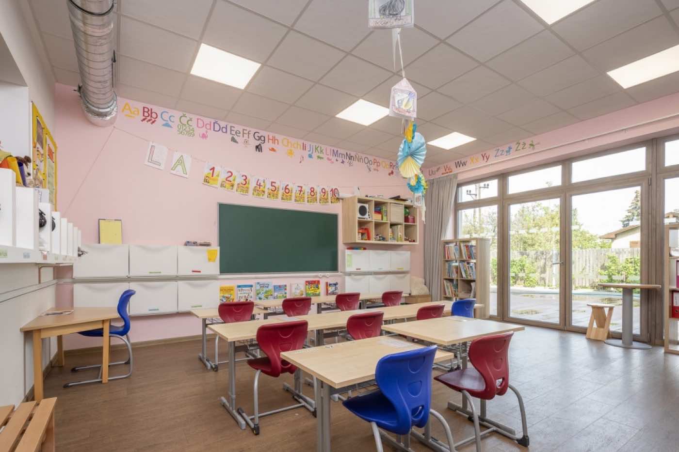 Světlá třída prvního stupně s dřevěnými stoly, modrými a červenými židlemi a zelenou tabulí